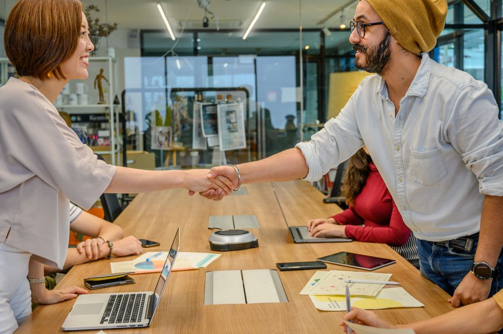 Professionals shaking hands across a meeting table with colleagues, laptops and project documents.